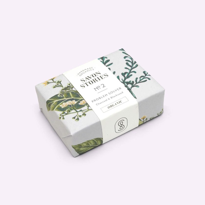 Rectangular bar of charcoal organic soap wrapped in paper printed with flowers and herbs on a grey background cream coloured sleeve with handmade organics, Savon Stories, le regulate charbon & Nigelle printed in black lettering
