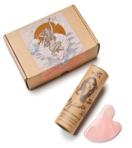 Rectangular cardboard box with goddess design next to cardboard tube with black print and image Aphrodite in cursive form and ORGANIC FACIAL OIL ROSEHIP DAMASK ROSE&GERANIUM 30 ml above a pink quartz heart shape face tool