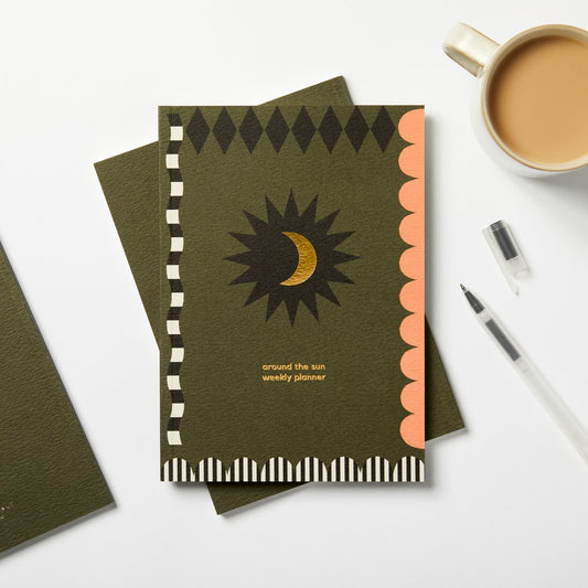 Dark olive green planner book with gold moon in a black star centred in the centre black diamond shapes along the top pink scallops down the right edge black and white striped scallops across the bottom and a black and white striped wavy line along the left edge around the sun planner is printed in gold foil lettering 