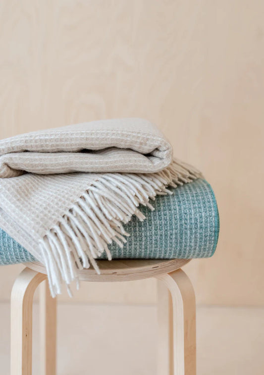 Large recycled wool sage green blue and cream waffle blanket with cream coloured tassels along the top and bottom edges folded on a stool