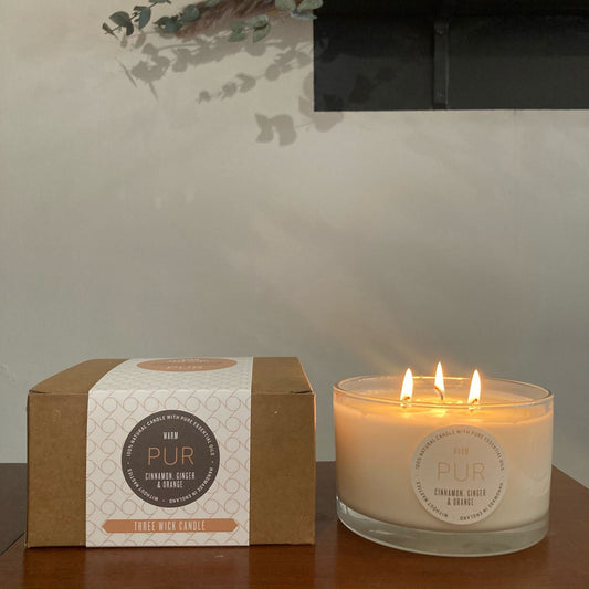 Candle in wide low glass jar with 3 wicks and flames round label with yellow print WARM PUR and grey print cinnamon ginger and orange 100% natural candle with pure essential oils handmade in England on a wooden table next to a craft box with cream coloured sleeve